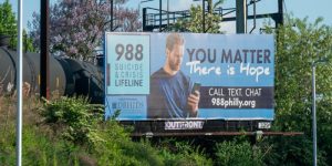 Ad promotion 988 Suicide and Crisis Lifeline as seen in Philadelphia (billboard) - ad depicts white man with beard looking at his phone. Text reads "You Matter - there is hope" - sign above a highway in daylight with a rail car behind it.